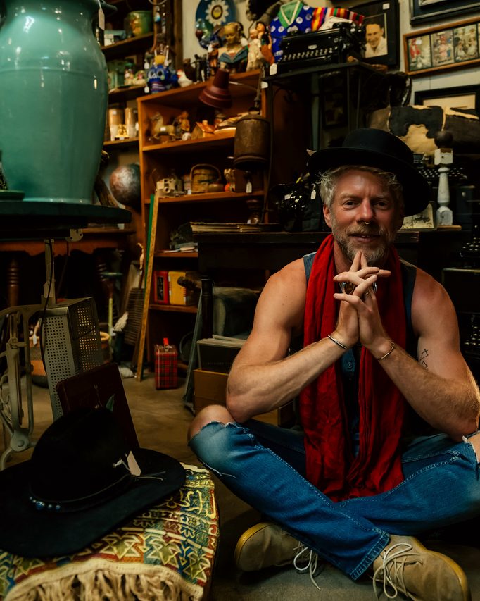 Seated cross-legged in an eclectic room, hands at the heart