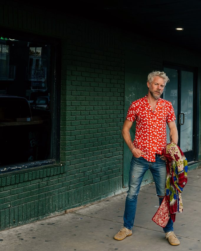 Standing on the street in a red patterned shirt against a green wall